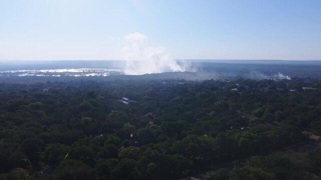 Aerial sweep over a treed Zimbabwean village shows the Zambezi River and Victoria Falls spray. Mist rises, smoke bands drift, sunlight glints in late afternoon haze.