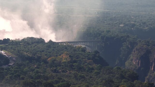 Aerial view shows Victoria Falls, the arched steel bridge, and Zambezi River. Mist drifts in warm late afternoon light over mopane woodland and village structures.
