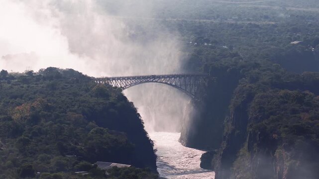 Aerial sunset view shows Victoria Falls in Zimbabwe, the arched Victoria Falls Bridge, and the Zambezi River rushing below, with mist drifting across the gorge.