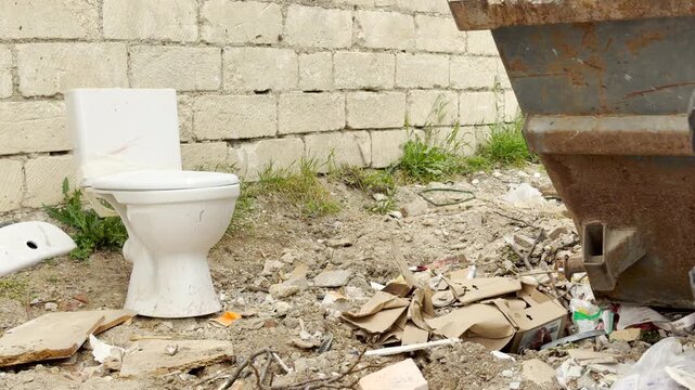 An abandoned ceramic toilet stands amidst a messy pile of garbage. The depressing scene shows scattered cardboard and debris on the dirt ground by a brick wall during a bright day.