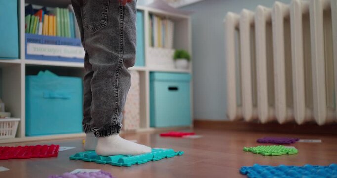 Toddler in socks stepping across colorful sensory puzzle mats on a wooden floor in a cozy playroom with storage cubes and a radiator