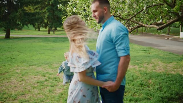 Family enjoys a peaceful afternoon in a lush green park. The father and daughter stand close, smiling and looking toward the running toddler. The child dashes joyfully toward them, arms outstretched