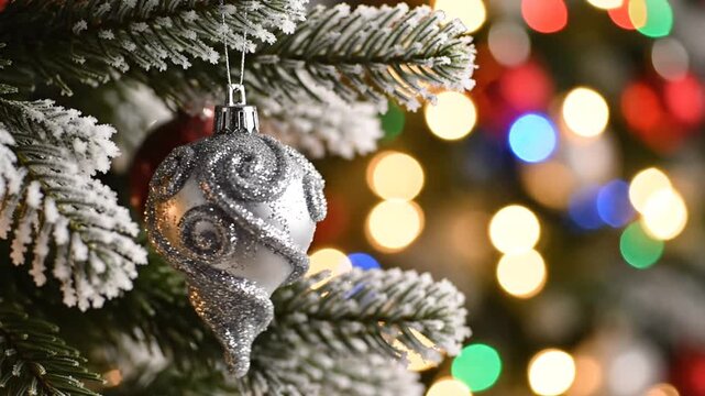 Close-up of a silver glitter ornament with embossed swirl patterns hanging on a frosted pine tree branch with blurred colorful bokeh holiday lights in the background.