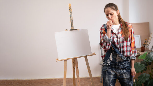 Unwell female artist coughing in fist during creative burnout or sickness concept. Contemporary art studio with blank white canvas mockup and clean copyspace for advertising text banner