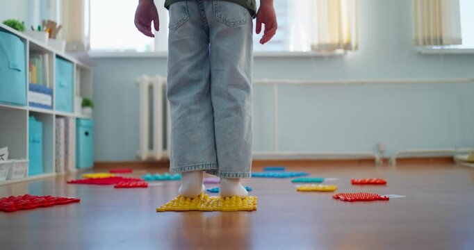 Child in socks walking across colorful sensory mats on a wooden floor in a bright playroom, practicing balance, coordination, and tactile exploration