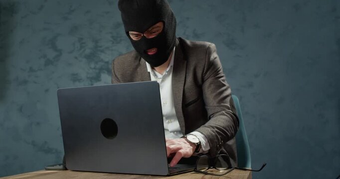 Masked scammer gestures angrily near laptop during failed online fraud attempt at desk. Man raises hands reacting emotionally while facing computer screen during scam