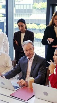 Team in an office huddles around laptops during a tense meeting, with one colleague thinking hard as others discuss data and strategy by the window.