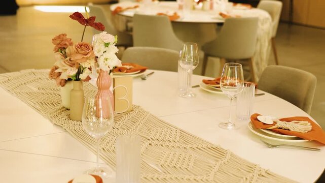 Wedding table number three lettering on an elegant dinner tablescape layout. High angle slow motion close up of a beautiful marriage banquet decoration with boho macrame runner and floral vases.