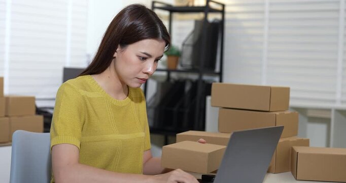 Asian businesswoman updating order list on laptop after scanning barcode, organizing e-commerce parcel boxes, managing inventory for delivery, home office small business logistics workflow.