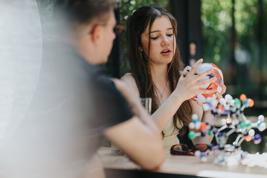 A teenage girl and classmate school friends examine a molecular model together. The students discuss the science model casually at a table outdoors.