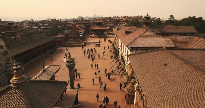 Aerial view across the wide brick-paved plaza of Bhaktapur Durbar Square, with tourists strolling between ancient temples and pagoda rooftops at sunset.