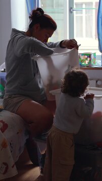 Mother organizing children toys into storage containers while toddler explores nearby during cleanup routine capturing parenting multitasking and authentic family home life