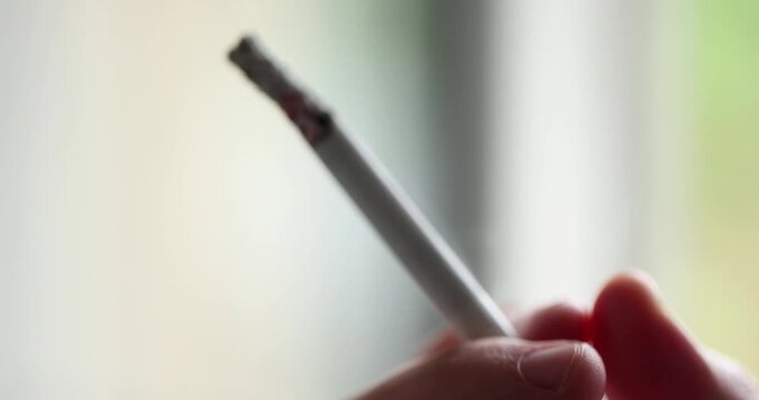 A lit cigarette is held between fingers, with ash forming at the tip while smoke gently rises in the soft lighting and gloomy place.