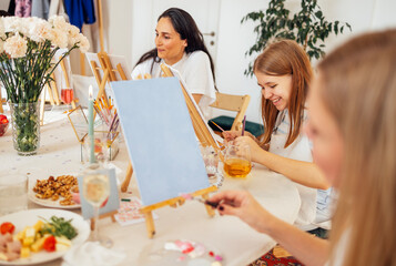 Group of women engaged in creative painting activity at a bright studio, with easels, colorful paints, and snacks on the table, fostering artistic expression and social interaction © Dasha Petrenko