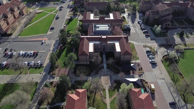 Symmetrical academic complex south of Colorado Ave anchors view landscaped paths, courtyards, parking areas connecting to neighborhood streets. Warm roof tiles, stone facade, trees, Boulder, CO