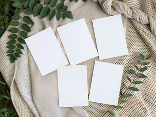 Fototapeta premium Five blank vertical cards mockup set on white knitted blanket. Empty stationary templates or wedding invitation collection with green leaves decoration outdoor.