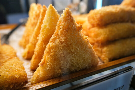 Crispy, jagged texture of the panko breadcrumbs covering a deep-fried, triangular risoles pastry resting on a tray. Relevant bakery merchandise, savory snack frying, cafe appetizers, party snacks