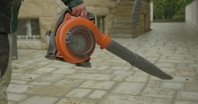 Person wearing green jacket and cargo trousers holding and operating an orange petrol leaf blower on a stone patio