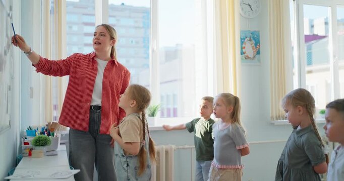 Teacher guiding young children at a classroom whiteboard during a bright interactive lesson, with students watching, drawing, and learning together