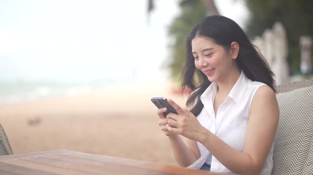 Asian woman using smartphone at tropical beach cafe, Smiling young female messaging, reading online content on mobile phone in summer vacation, online travel booking, Travel and communication concept