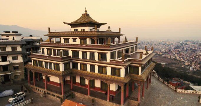 Aerial drone shot of the ornate Chovar Gumba Buddhist monastery in Nepal, with its multi-tiered pagoda roof and golden ornaments overlooking the hazy Kathmandu cityscape at sunset.