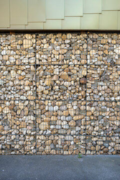 A gabion wall filled with rounded stone and pebble texture sits below a plain facade, its metal structure meeting pavement in an urban edge