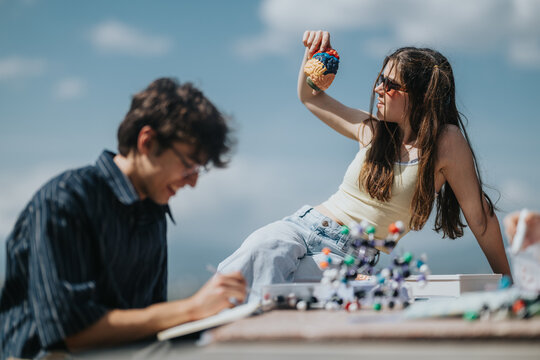 A teenage girl and boy study science outdoors with a colorful anatomical brain model and molecular models on a table. They interact with educational props while taking notes and examining models.