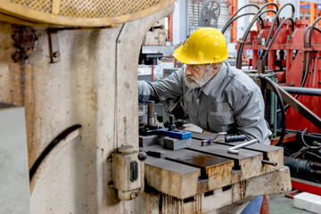 Fototapeta premium Senior male worker with safety clothing, helmet and gloves performs maintenance and repairs on machinery used in factory production. Elderly technician working and inspecting a manufacturing machine.