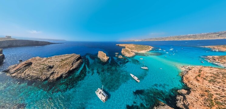 Wide, aerial, panoramic photo of the rocky coast of Comino Island in Malta, with a view of the Blue Lagoon and the yachts moored in the bay.