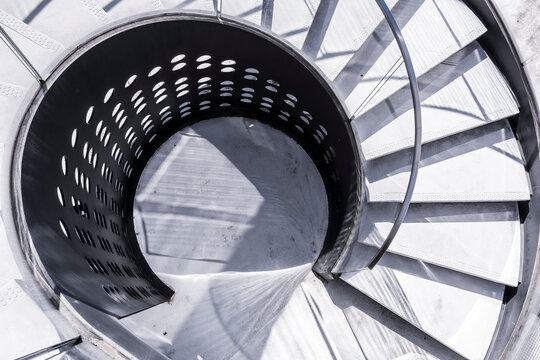 A spiral staircase curls around a circular void, where concrete steps, shadow pattern, handrail, and architectural geometry create a graphic view