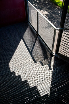 Perforated metal stairs fall into shadow near a railing, where sunlight, pattern, texture, and urban architecture sharpen the exterior geometry