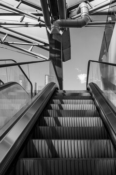 A monochrome escalator rises between metal rails, where stairs, handrail, machinery, architecture, and shadow turn transit movement into graphic rhythm
