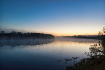 Fototapeta premium A river reflects the sky at dawn, with mist hovering over the water and trees bordering the edges. The sun begins to rise, lighting the scene softly