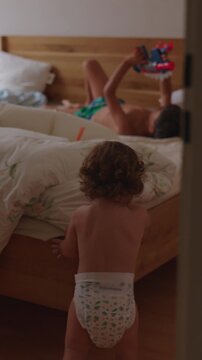 Toddler walking through bedroom toward older sibling playing with toy blasters on bed during lively family moment expressing curiosity interaction and authentic childhood home life