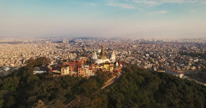 Aerial wide shot of the forested Swayambhunath hilltop temple complex perched above the misty, expansive Kathmandu Valley at dawn.