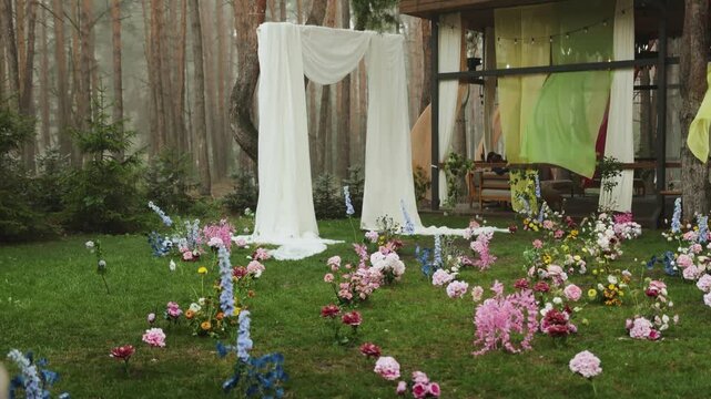 At wedding party decor raining on flowers in green grass lawn near marriage arch in forest 4K. Beautiful cinematic outdoor ceremony setup during rainy day with ground floral arrangements, water drops