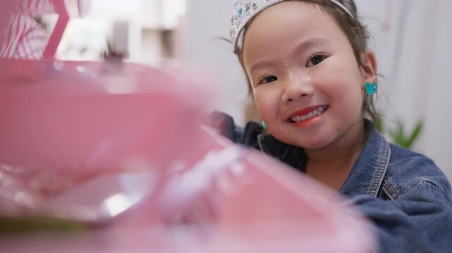 A happy young girl wearing a princess tiara and makeup smiles at the camera while playing dress-up, expressing her childhood imagination and joyful creativity at home.