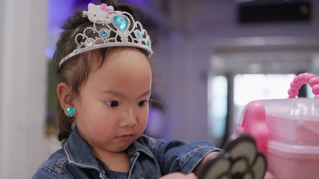 A young toddler girl wearing a tiara plays with makeup, exploring creativity and self-expression during her childhood development in a close-up portrait.