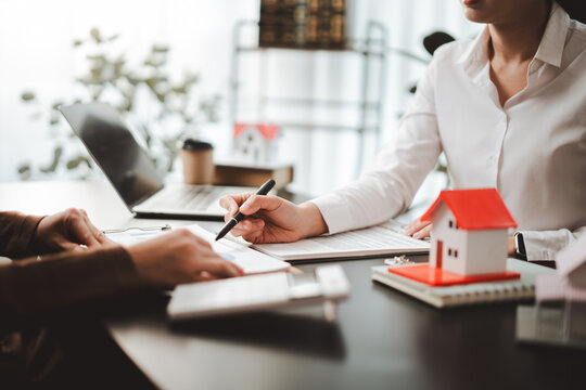 Real estate agent and client at a desk reviewing mortgage documents and a small model house, discussing contract details, financing options and homeownership plans