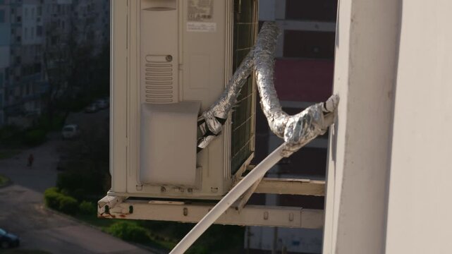 White outdoor air conditioning compressor unit sits mounted on a rusty metal bracket against a building facade. Foil-wrapped pipes connect to the system. Concept of urban climate control.