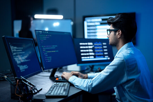 Young male software engineer writing program code on multiple desktop screens while his colleague working in the background in a modern office.