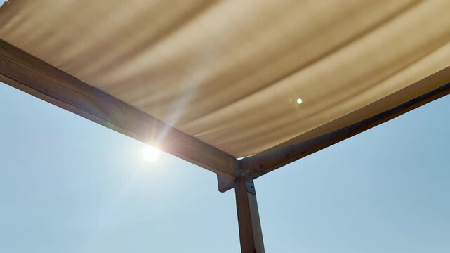 Warm sunlight shines brightly past the corner of a sturdy wooden pergola. A soft beige canvas shade overhead ripples gently, creating a serene and uplifting summer atmosphere.