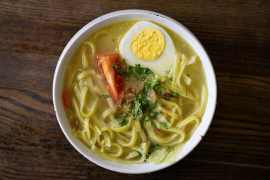 A bowl of Javanese noodle soup, topped with sliced egg, celery leaves, chopped tomato, and a thick broth, looks delicious. 