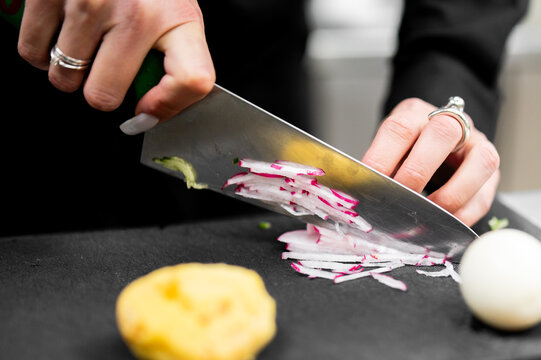 A chef slicing red radishes on a black cutting board with a professional knife, close up.