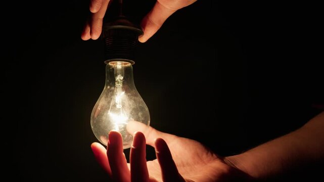 Hands screw a brightly glowing incandescent light bulb into an overhead socket against a pitch-black background. Concept of sudden inspiration, creative ideas, and electricity.