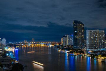 Obraz premium Stunning panoramic view of the Bangkok city skyline at night with modern skyscrapers reflecting on the Chao Phraya River under a cloudy sky in Thailand. Perfect for travel concepts.