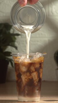 Close-up of making a takeaway iced coffee cocktail. Milk is poured from a glass jug into a plastic cup filled with layers of condensed milk, ice cubes, caramel, and coffee