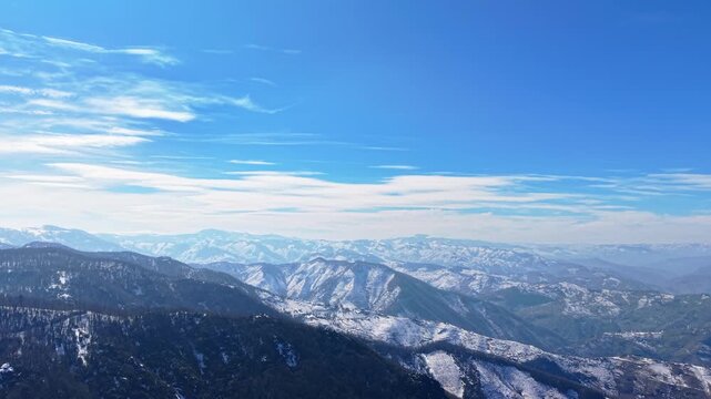 A cinematic drone shot starting at mountain ridge level, facing directly forward toward snow-covered peaks. The camera remains steady in orientation while slowly rising upward, gradually revealing mor