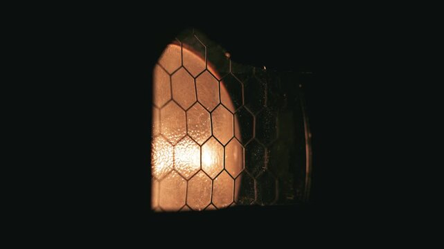 Macro shot of an industrial stage light fixture. Warm orange tungsten light glows through a distinct hexagonal honeycomb mesh against a dark background with subtle lateral camera movement