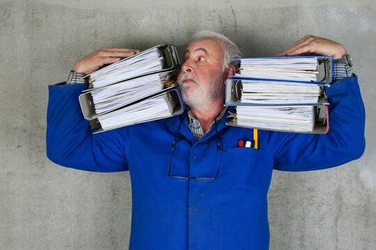 Stressed senior craftsman carrying heavy folders, symbolizing excessive bureaucracy, paperwork overload and administrative pressure on small business and trades in Germany and Europe.